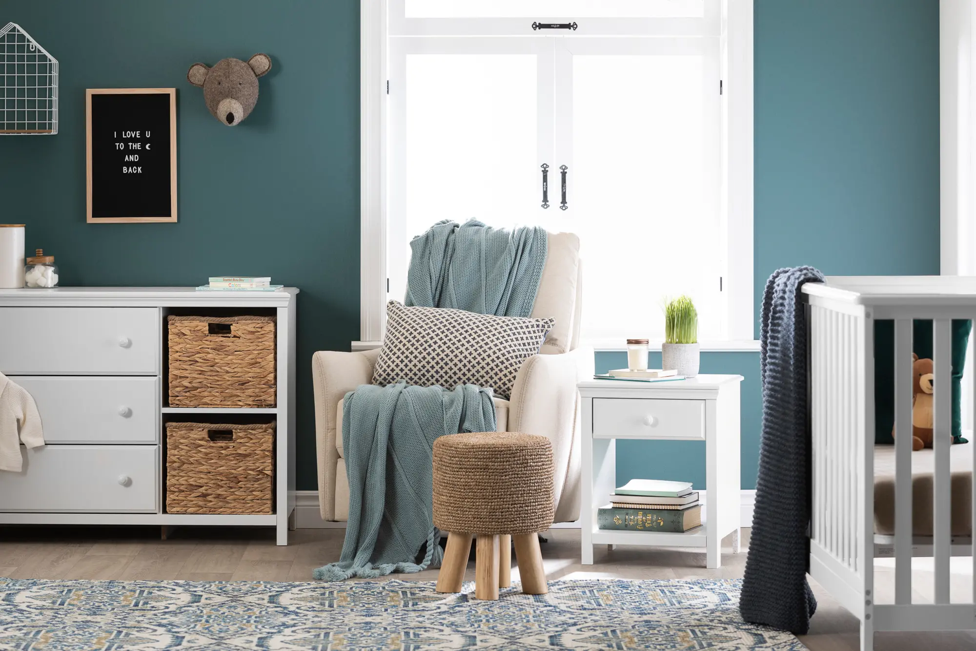 Child bedroom with teal walls, white dresser and nightstand, crib, and decorative rug.